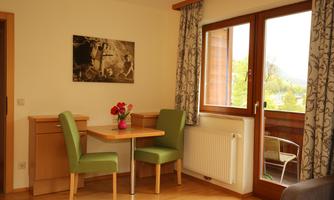 A cozy dining area with a table and two chairs. Through the window, you can see a balcony and plenty of daylight.