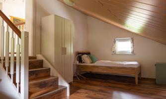 A bright bedroom under the roof with a single bed and a wardrobe. The floor is made of wood and there is a staircase that leads down.