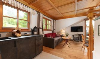 A cozy living room with wooden paneling and large windows. It includes a kitchen, a sofa, and a television, surrounded by an inviting atmosphere.