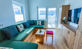 A modern living room with a green couch and a wooden table. Large windows provide plenty of daylight and a view of the surroundings.