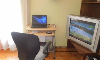 A workspace with a laptop on a table and a television in the background. The chair is aligned with the table, next to it is a window with natural light.
