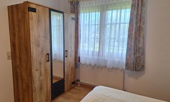 A bedroom with a wooden wardrobe and a large window. The walls are light and the windows have curtains.
