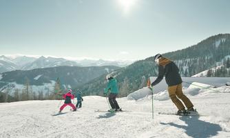 A group of skiers is skiing on a snowy slope. In the background, majestic mountains and a bright blue sky can be seen.