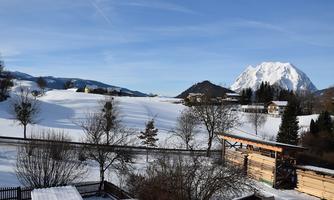 A snowy landscape with mountains in the background and a clear blue sky. There are scattered trees and buildings that complement the tranquil scenery.