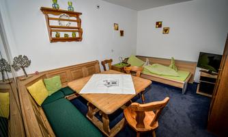 A cozy living area with a wooden table and benches in warm colors. Decorative pictures hang on the walls, and a television is placed in the corner.
