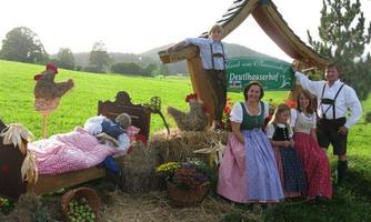 A traditional Bavarian scene with people in traditional costumes. In the background, a deck chair and decorative chickens can be seen, as well as a green meadow.