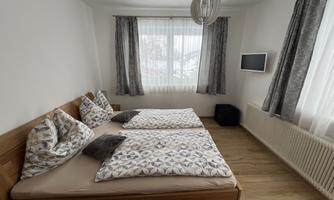 A cozy bedroom with two single beds and modern pillows. The room has large windows and a wall with a television.