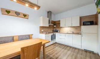 A modern kitchen with wooden shelves and white kitchen furniture. A wooden table with chairs is in the foreground.