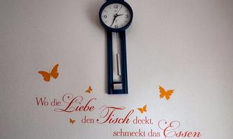 A wall clock in blue color hangs on a white wall. Below it is a saying about love and food, surrounded by orange butterflies.