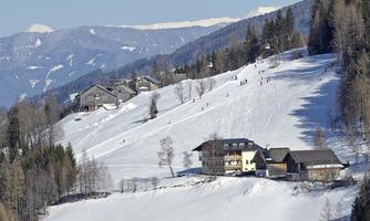 A picturesque winter landscape with snow-covered hills and skiers. In the foreground, several wooden houses and trees can be seen.