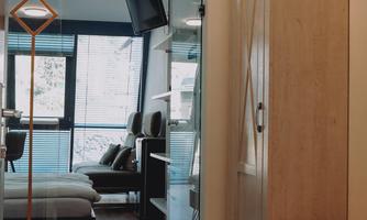 A modern interior with wooden flooring and a large window. On the left side, a wardrobe and a comfortable seating area are visible.