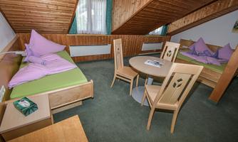 A cozy room with a wooden ceiling, two beds, and a round table. The walls are clad in wood, and the windows let in plenty of light.
