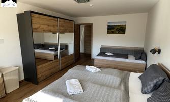 A modern bedroom with a large double bed and a single bed. The room features light wood and large windows for plenty of daylight.