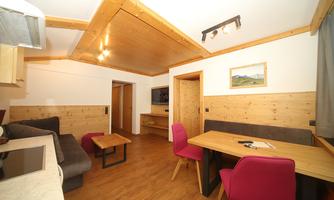 A cozy living room with wooden paneling and modern furniture. There is a dining area with pink chairs and a comfortable sofa area.