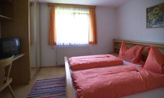 A simple bedroom with a double bed and orange pillows. There is a wardrobe, a chair, and a window with curtains.