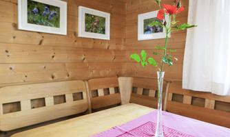A cozy room with wooden walls and two wooden benches. On the table stands a single red flower in a glass vase.