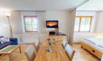 A bright and cozy living room with a dining table and a sofa. On the wall, there is a television, and the windows let in plenty of light.