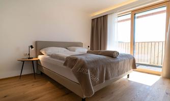 A modern bedroom with a large bed and wooden flooring. Large windows provide plenty of daylight.