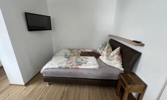 A cozy room with a bed, decorated with colorful pillows. A television is hanging on the wall, and the floor is covered with wooden planks.