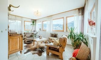 A bright living room with large windows and a view of nature. It is cozily furnished with plants, a coffee table, and a stylish armchair.