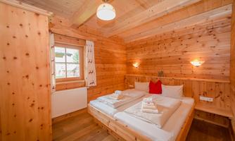 A cozy wooden room with a double bed and white bedding. The walls and ceiling are made of wood, and the window lets in plenty of daylight.