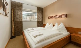 A modern bedroom with a double bed and white bedcovers. The walls are painted in soft colors, and a large window lets in plenty of daylight.