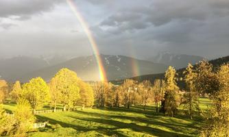 A rainbow appears above a picturesque landscape with green meadows and trees. In the background, mountains and a cloudy sky can be seen.