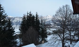 A winter landscape with snow-covered mountains and trees. The sky is clear and slightly blue.