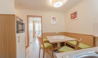 A bright kitchen with a dining area and modern furniture. A table with green chairs and a large window opening creates an inviting atmosphere.