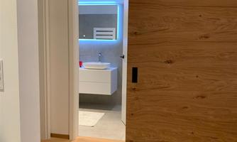 A cozy entrance area with a wooden sliding door. In the background, a modernly designed bathroom with blue lighting is visible.