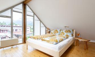 A cozy bedroom with a large window and a wooden floor. The bed is decorated with colorful bedspreads and pillows.