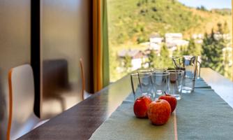 A well-set table with red apples and glasses stands in front of a window overlooking a green landscape. The atmosphere is bright and inviting.