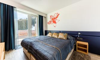 A modern bedroom with a large bed and a blooming wall decoration. The windows provide access to a balcony overlooking nature.
