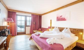 A cozy room with a large double bed and wooden decor. The windows provide a view outside and the walls are painted in warm colors.