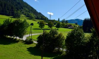 A picturesque landscape with green meadows and trees. In the background, gentle hills and a blue sky are visible.