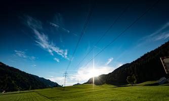 A vast green field under a clear blue sky. The sun is low and shines through the mountains.