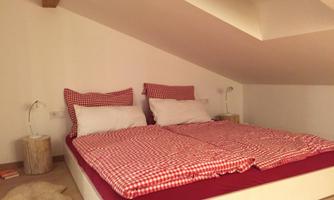 A cozy bedroom with a double bed and red-and-white checked bedcovers. The walls are simple and bright, with a sloping ceiling and minimalistic furniture.