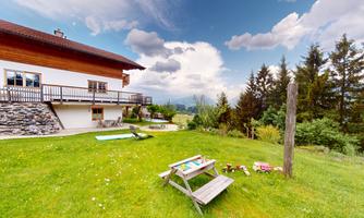 A cozy meadow with a wooden house in the background. There are garden furniture, toys, and a lovely view of the surrounding nature.