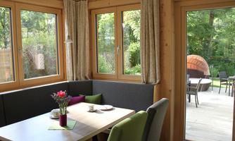 A cozy dining area with wooden decor and large windows that offer a view of the garden. Flowers and dishes are on the table, while a terrace with seating can be seen outside.