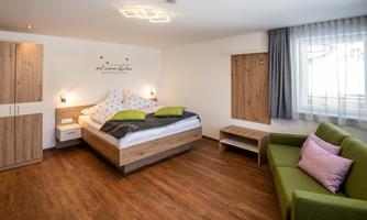 A modern bedroom with a large bed, a green sofa, and wooden details. Large windows let in plenty of daylight.