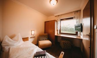 A simple room with a bed, a television, and a window. The walls are light, and there is a cozy armchair.