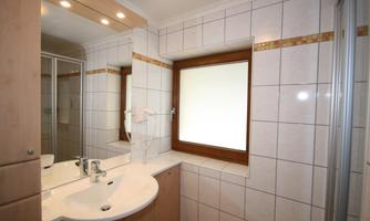 A bright bathroom with tiles and a large window. There is a modern shower and a sink with two faucets.