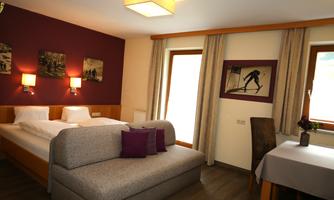 A cozy hotel room with a double bed and a sofa. The walls are painted in warm colors, and there are large windows with a view of the outside.