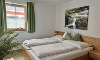 A cozy bedroom with a double bed and green curtains. A picture of a waterfall hangs on the wall, and a plant is standing on the floor.