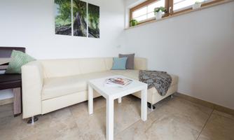 A modern living room with a beige sofa and a white table. Pictures of trees hang on the wall, and there are plants on the window sills.
