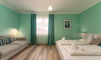 A modern room in soft turquoise tones with a cozy sofa and a window. The bed is neatly made and the curtains are open, allowing plenty of light in.