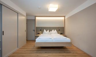 A modern bedroom with a double bed and light wood details. The walls are painted in neutral colors and there is a large wardrobe.