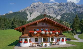 A traditional chalet with blooming flower boxes amidst mountains. The green meadow and clear sky give the scene a peaceful atmosphere.