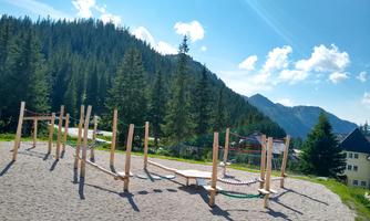 A wooden playground with various climbing elements is situated on a sandy ground. In the background, tall trees and mountains can be seen under a clear sky.