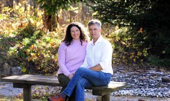 A couple is sitting on a bench in a sunny wooded area. In the background, colorful leaves and a small gravel path are visible.